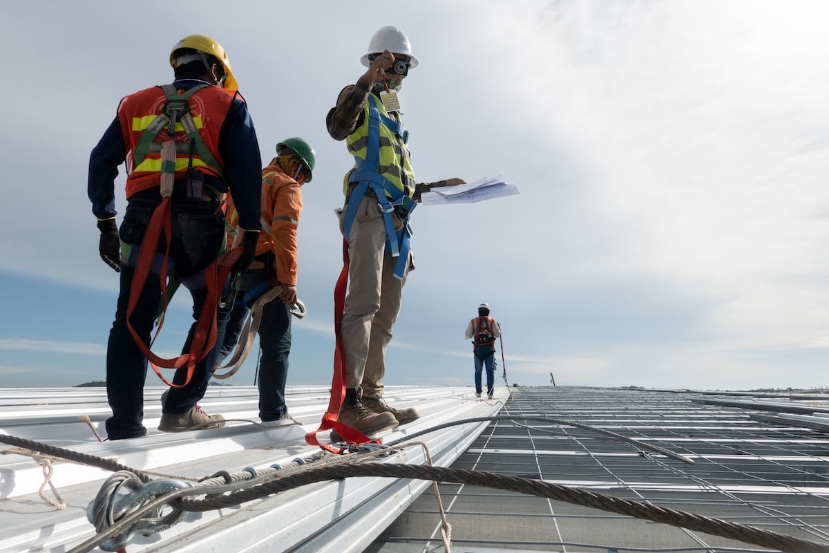 Four commercial roofing contractors wearing safety gear and harnesses stand and walk on a metal rooftop under a blue sky, with one person holding blueprints and observing the site.