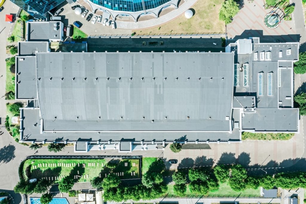 Aerial view of a large rectangular building with a gray roof, likely maintained by commercial roofing contractors, surrounded by trees, grass, and walkways. Several smaller structures and parked vehicles are visible nearby.