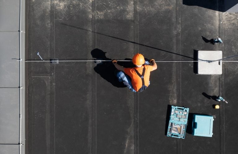 A worker in an orange shirt and hard hat kneels on a flat roof, performing flat roof repair with a metal cable. Tools and a blue toolbox are nearby on the rooftop.