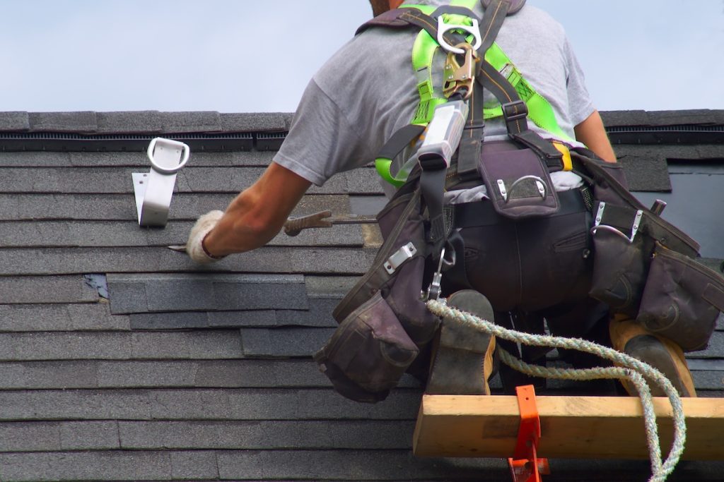 A construction worker wearing safety gear installs or repairs asphalt shingles on a roof,