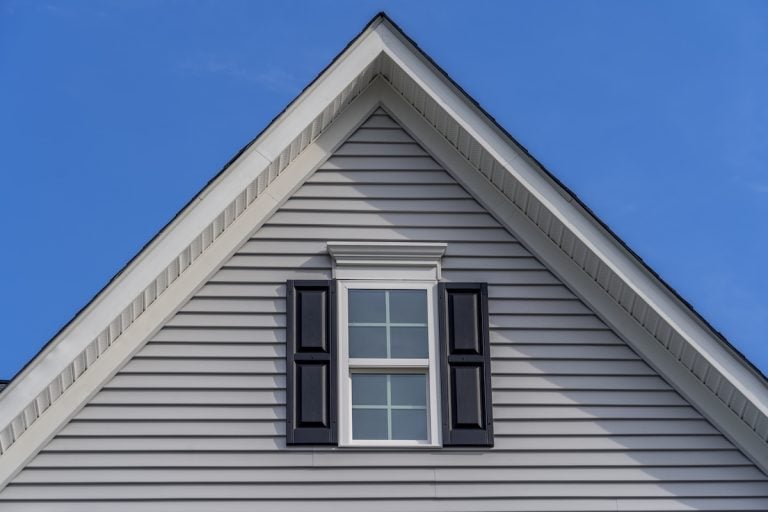 Close-up of the gable of a house with light gray siding, featuring a double-hung window with white trim and black shutters. The gable showcases a clean minimum roof pitch, set against a clear blue sky.