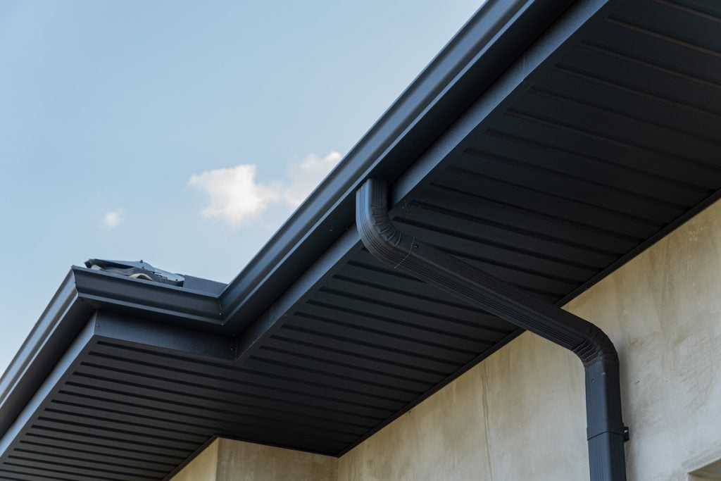 Close-up view of a modern black gutter system, one of the popular types of gutters, attached to the roof of a beige building, with a blue sky and a few clouds in the background.