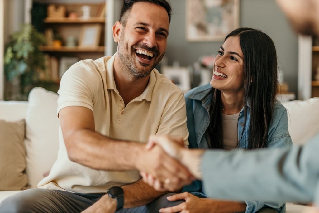 A smiling man and woman sit on a couch, with the man shaking hands with someone off-camera—likely after a positive meeting about roofing insurance claims for storm damage. Both appear happy and relaxed in their home setting.