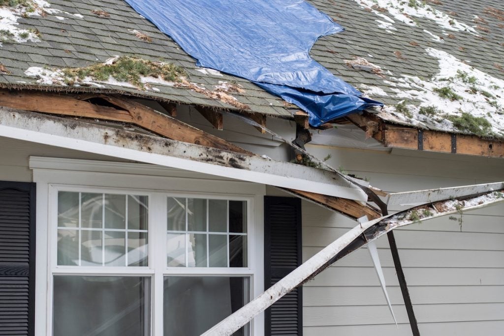 A house with roof damage, exposed wooden beams, and a blue tarp covers part of the roof. 