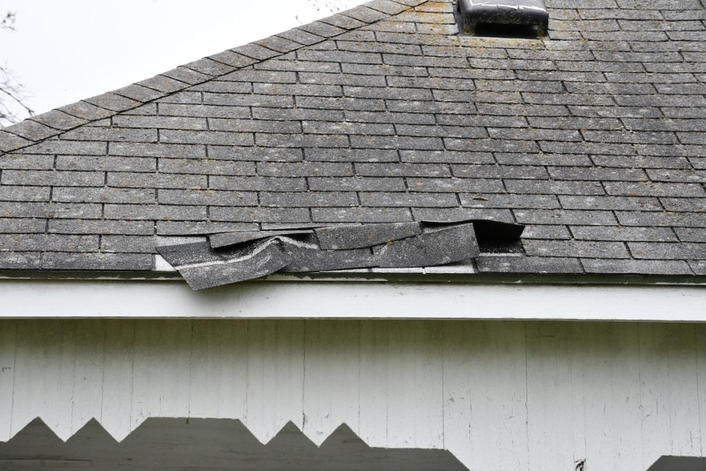 Damaged gray roof shingles, lifted and bent from hail and wind damage to the roof, expose the underlying structure near the edge of a white house with decorative trim under the eaves.
