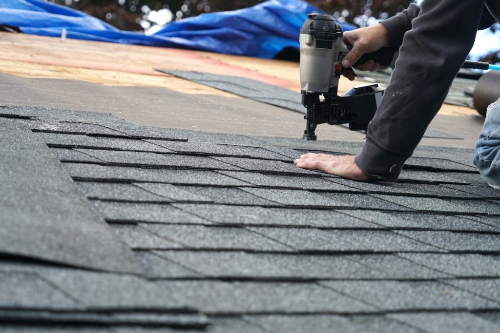 A person uses a nail gun to install asphalt shingles on a roof, demonstrating an important step from the roof maintenance checklist, with rows of partially completed shingles and construction materials visible in the background.