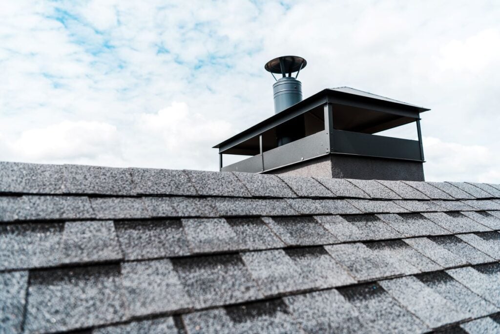 chimney rain cap Close-up of gray asphalt roof shingles with a black metal chimney rain cap vent extending above the roofline against a partly cloudy sky.