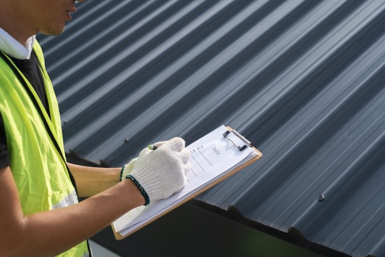 A person wearing a yellow safety vest and white gloves is holding a clipboard and writing notes while inspecting a dark metal roof, carefully following a roof maintenance checklist.
