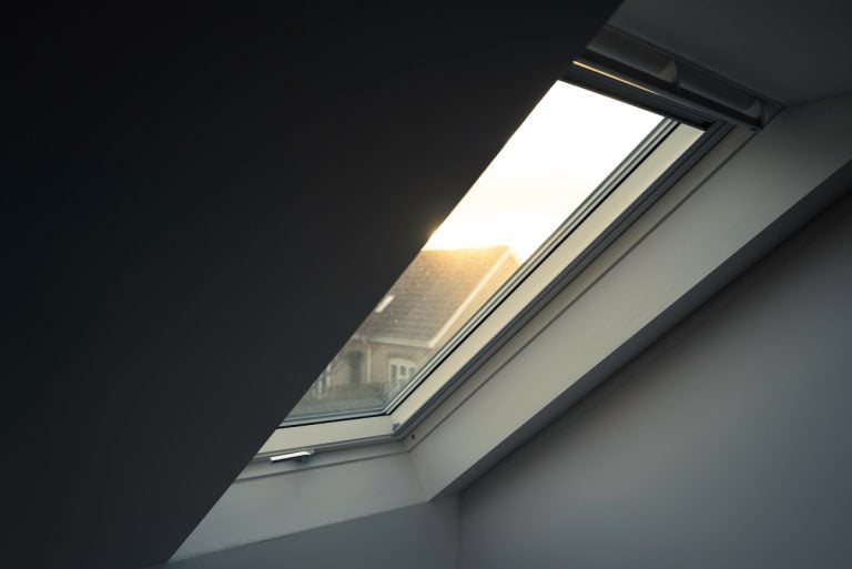 A slanted skylight window, one of the popular types of skylights, lets soft sunlight into a modern attic, with part of a neighboring house’s roof visible outside against a bright sky.