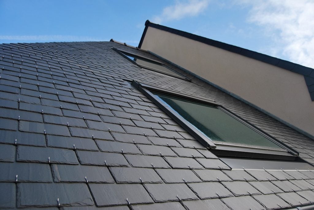 Close-up view of a modern sloped roof covered with dark slate tiles, featuring two rectangular skylight windows—showcasing popular types of skylights—set against a partly cloudy blue sky.