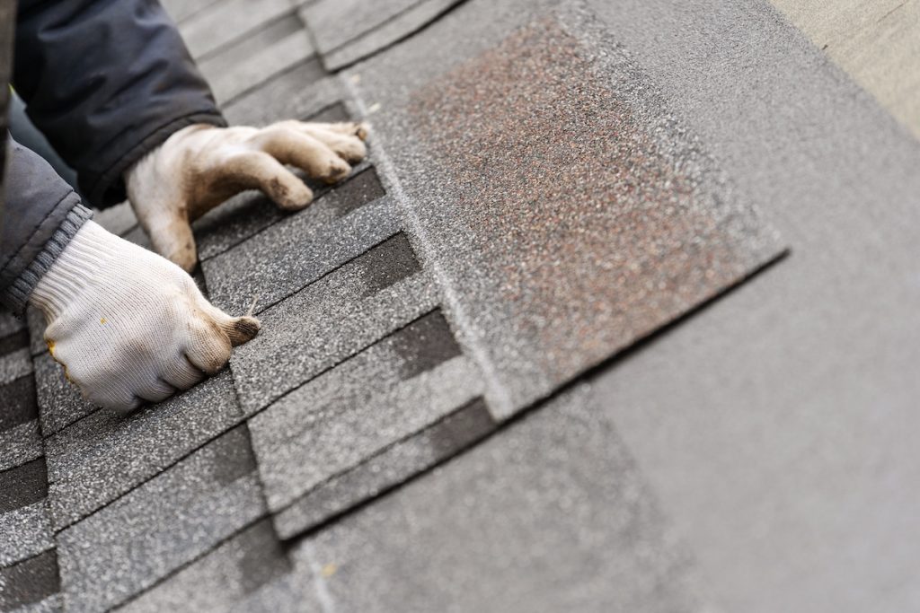 Close up and real photo of skilled workman in special protective work wear installing asphalt or bitumen tile on top of the roof under construction house