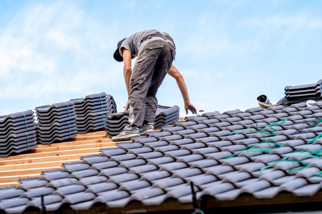 A worker wearing dark clothing is installing black roof tiles on a wooden roof frame 