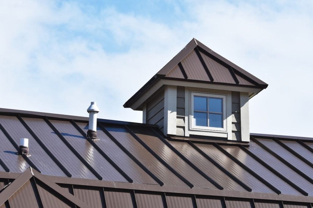 A brown metal roof with a small dormer window and two metal vent pipes, set against a partly cloudy blue sky
