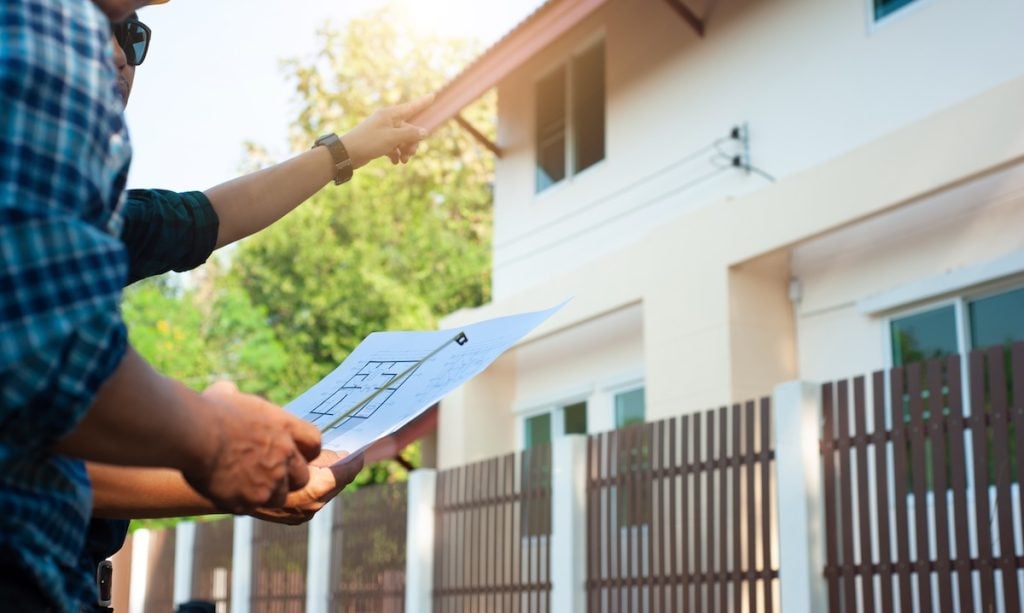 Two people stand outside a house with a cedar shake roof
