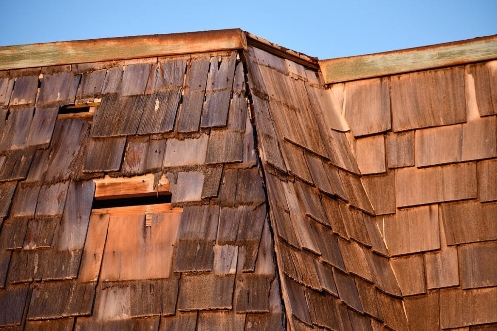 A close-up view of a weathered cedar shake roof with visible signs of damage, including missing and warped shingles, under a clear blue sky.
