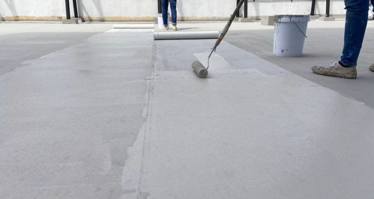 A person uses a paint roller to apply a white commercial roof coating on a flat concrete surface