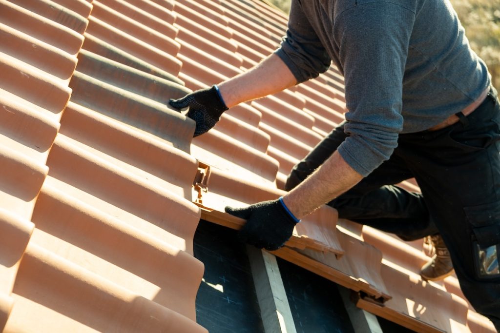 Closeup of worker hands installing yellow ceramic roofing tiles mounted on wooden boards covering residential building roof under construction.