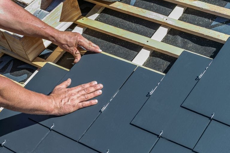 Slate roof repair A person’s hands installing dark rectangular roof tiles onto a wooden roof frame, securing them in place with metal hooks.