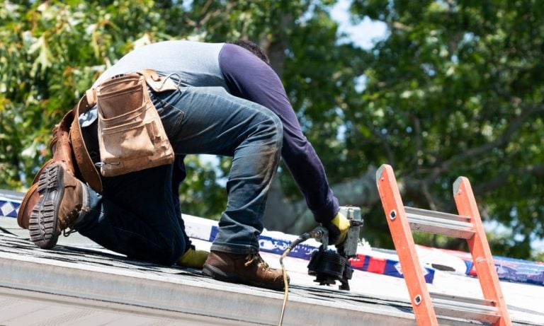 A person wearing work boots and a tool belt kneels on a roof, using a power tool. A red ladder leans against the roof, and trees are visible in the background.