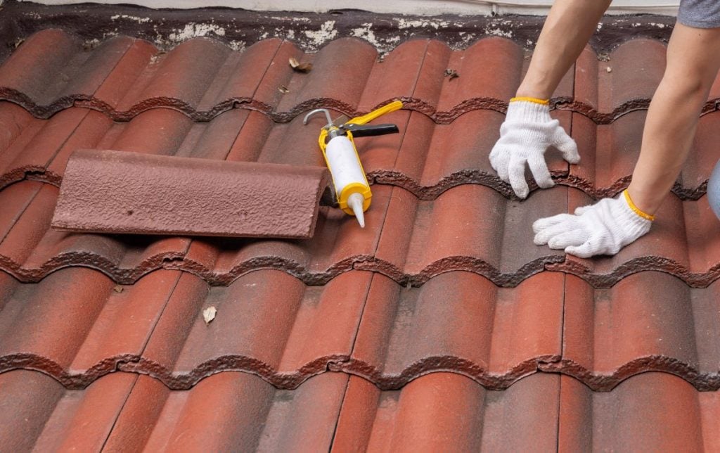 Person wearing white gloves repairs a red tile roof, with a caulking gun, tile roof sealer