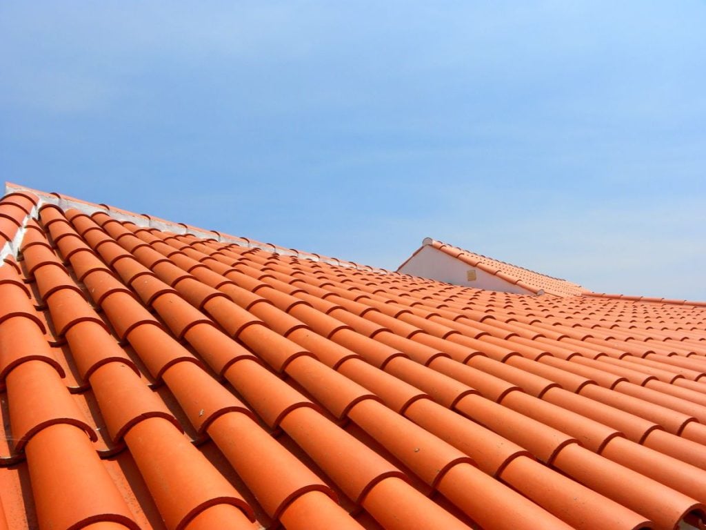 A close-up view of red clay roof tiles on sloped rooftops under a clear blue sky. The tiles are neatly arranged in rows, creating a pattern across the roofs.