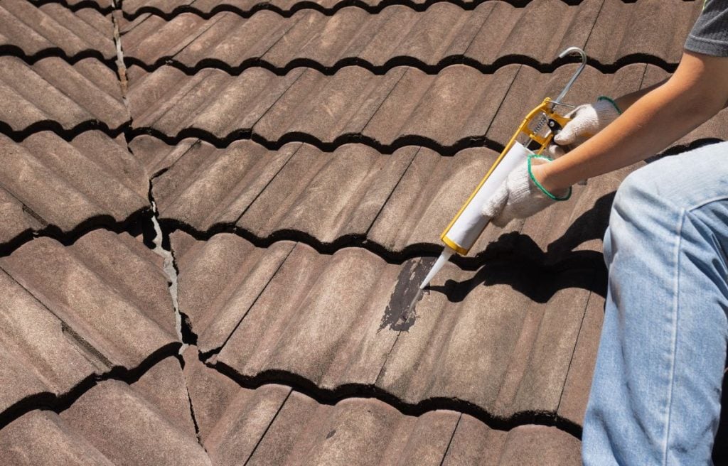 Worker man using silicone sealant adhesive to fix crack of the old tile roof.
