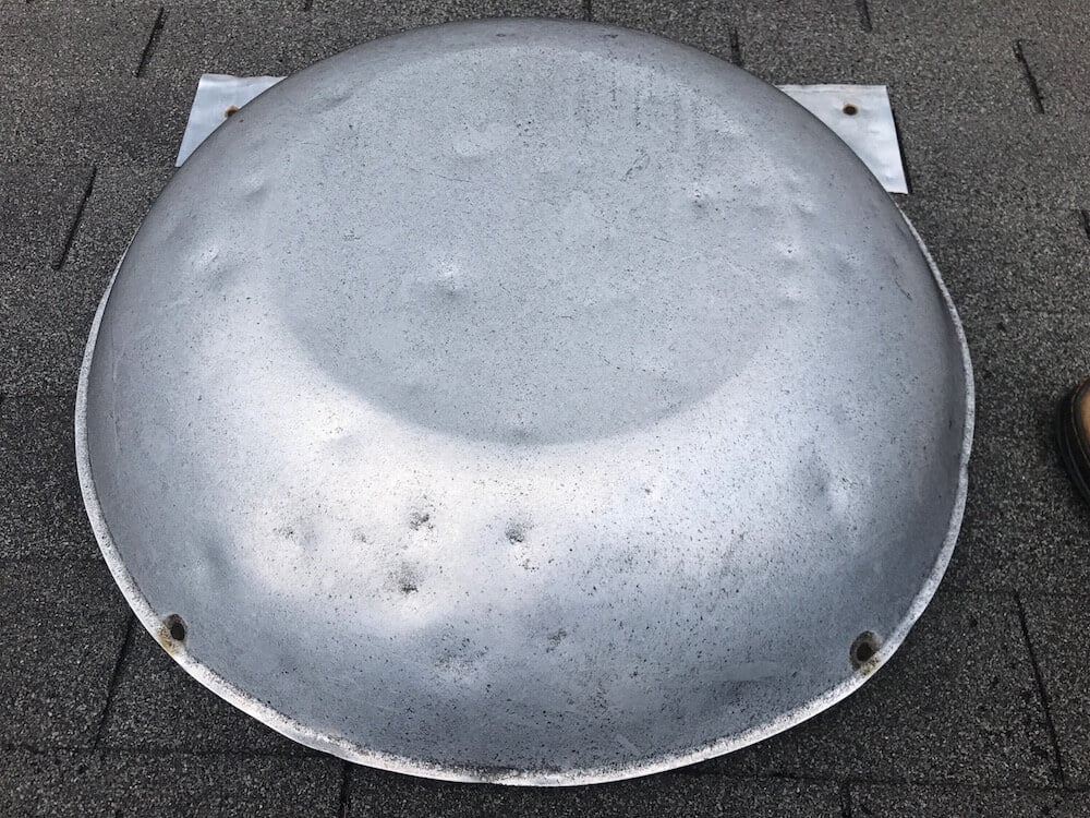 A large, round, metallic dome with small dents and three holes near the edge rests on a dark shingled surface