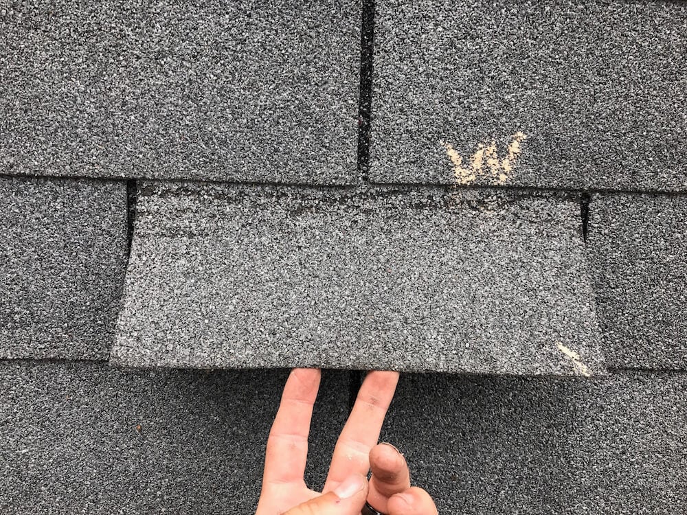A close-up of a hand lifting an asphalt roof shingle, wind damage