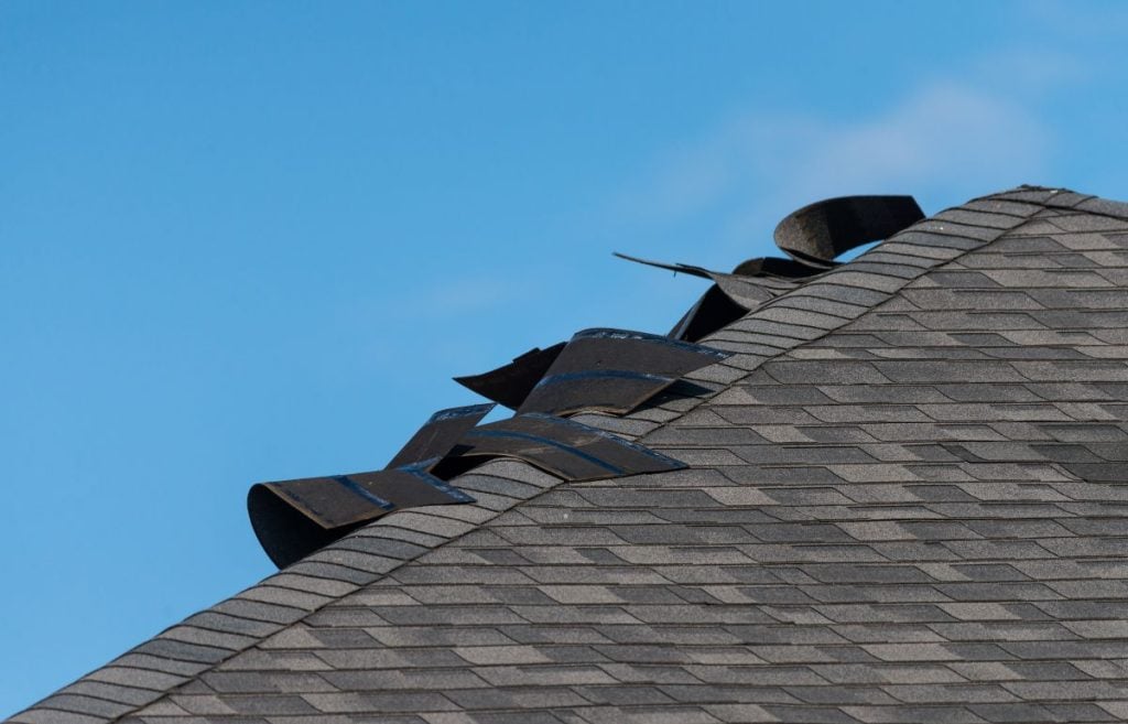 Close-up of a house roof with several asphalt shingles lifted and damaged