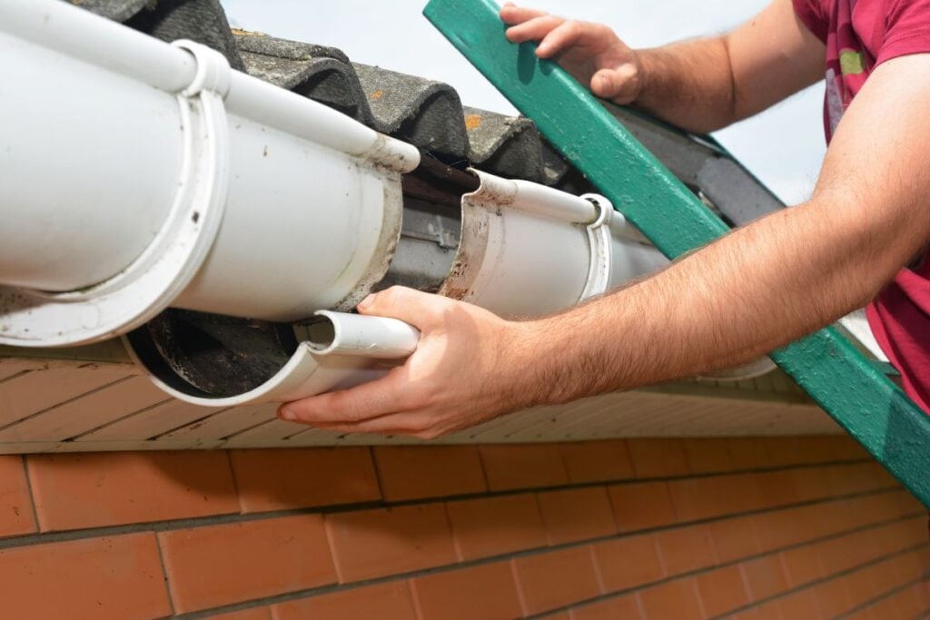 seamless gutters person standing on a ladder repairs or replaces a white plastic rain gutter attached to the edge of a sloped roof on a brick building.