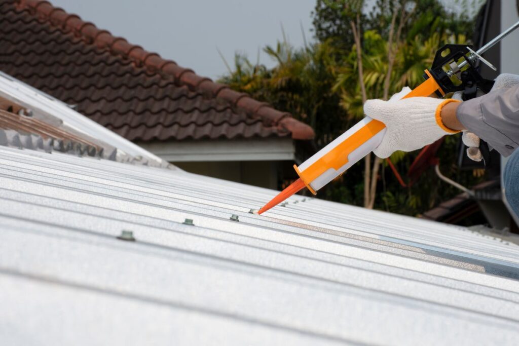 metal roof leak repair sealant person wearing white gloves applies sealant with a caulking gun to fasteners on a white metal roof, with a brown tiled roof and trees visible in the background.