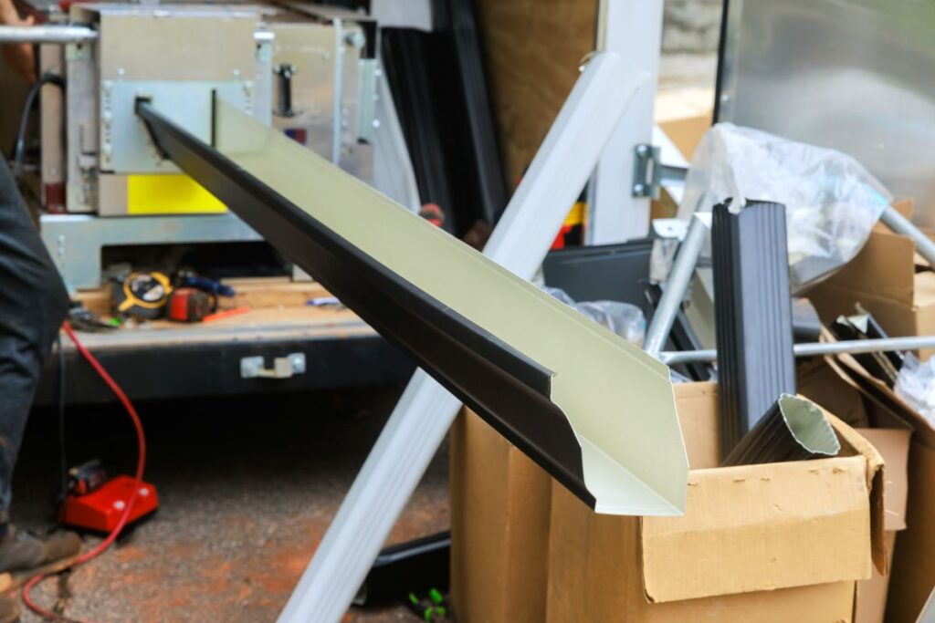 Long metal rain gutter being formed by a machine mounted on a work truck, with various construction materials and tools, including cardboard boxes and pipes, scattered nearby.