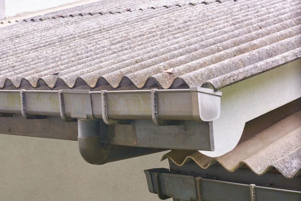 box gutters Close-up of a corrugated asbestos roof with a metal rain gutter and downspout attached to the edge of a residential building. The gutter appears weathered, and a few leaves are visible on the roof.