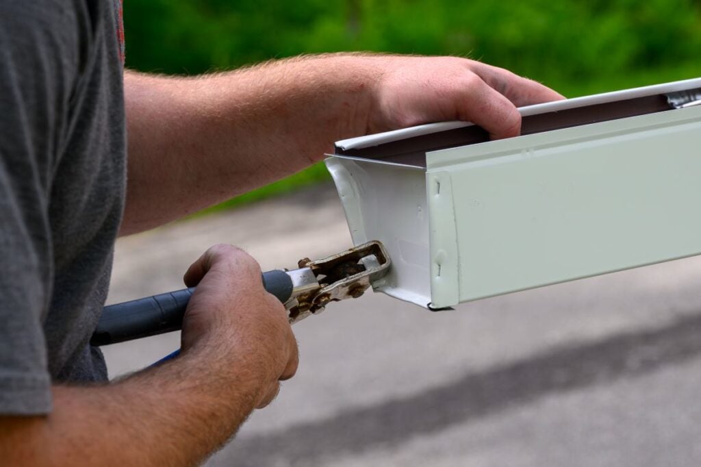 box gutters a person uses metal pliers to crimp the end of a white rain gutter, preparing it for installation. The background is slightly blurred, showing greenery and pavement.