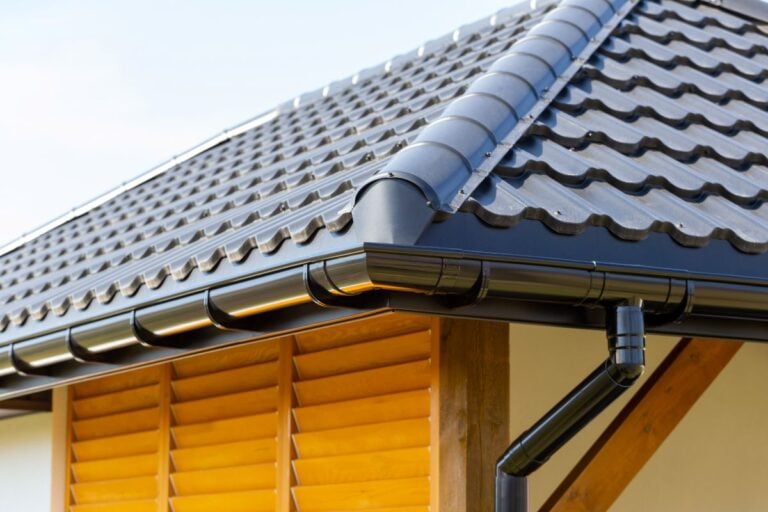 seamless gutters Close-up of a house corner with a black metal tile roof, black rain gutter system, and wooden louvered window shutters beneath the eaves, all against a clear sky background.
