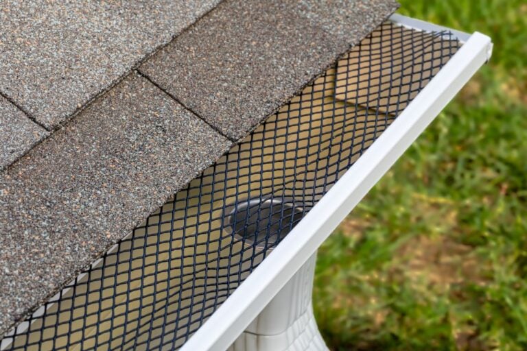 gutter screens Close-up of a house roof corner showing gray shingles, a white rain gutter, and a black plastic mesh gutter guard over the gutter, with green grass visible in the background.