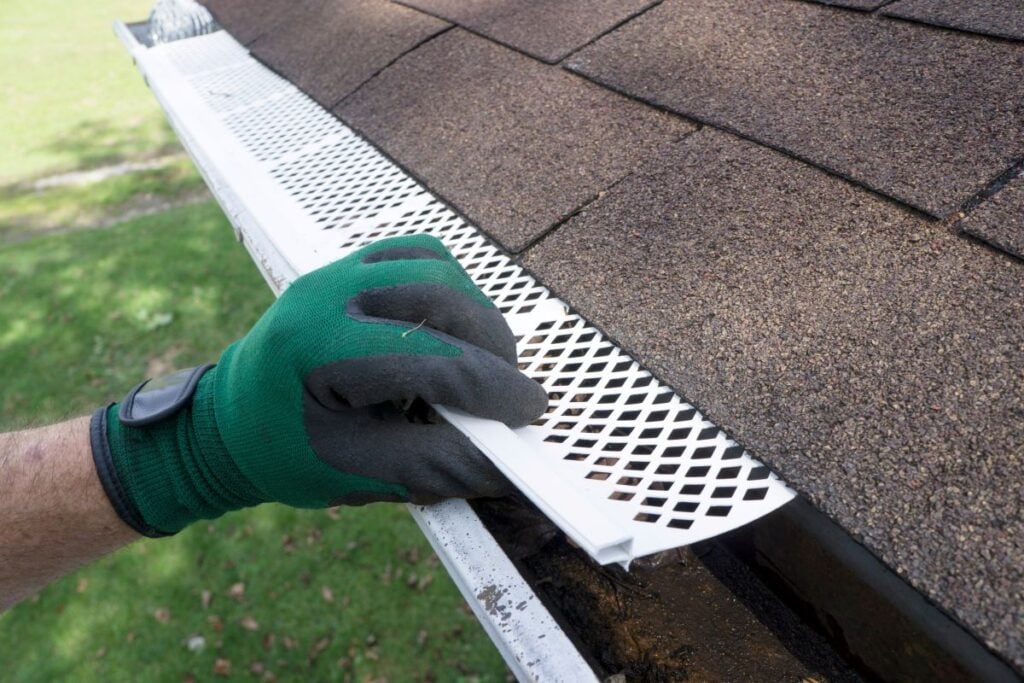 gutter screens a person wearing a green glove installs a white plastic gutter guard on the edge of a shingled roof, helping to keep debris out of the gutter below.