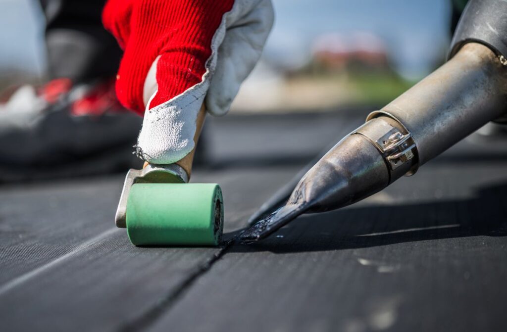 flat roof sealant close-up of a worker using a heat gun and a green roller tool to seal seams on a black waterproof membrane, wearing a red and white protective glove.