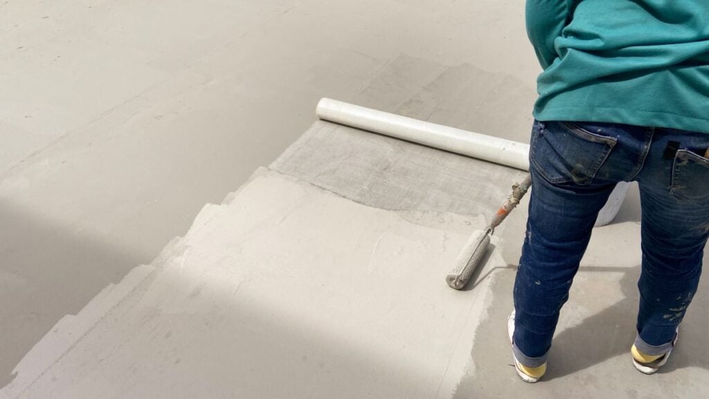 flat roof sealant person in jeans and a teal shirt uses a paint roller to apply a light gray coating over a surface, partially covering a piece of mesh material. The person is standing on the right side of the image.