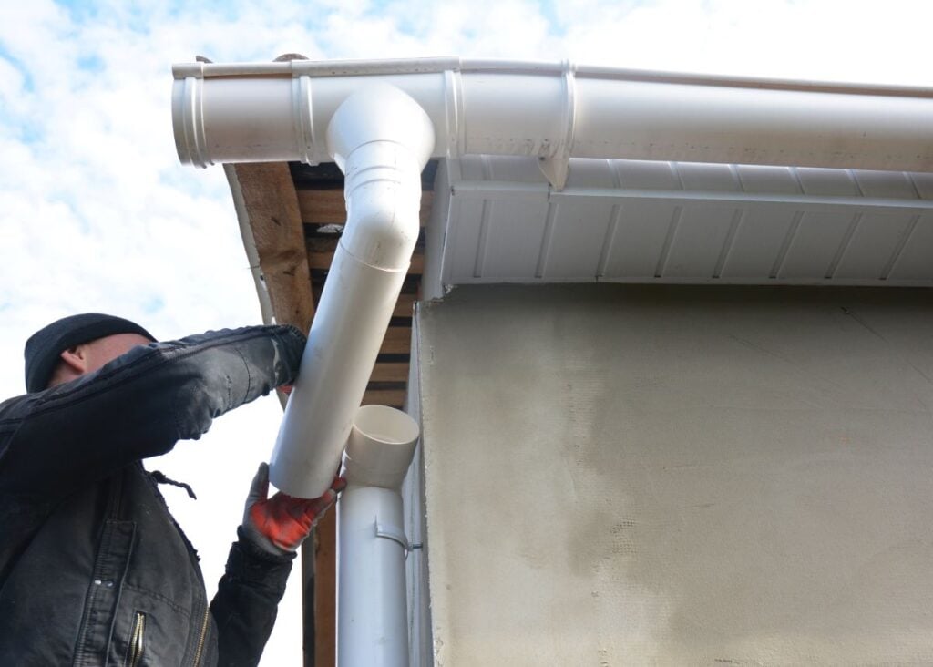 seamless gutters A worker wearing gloves installs a white rain gutter and downspout system on the edge of a building’s roof. The sky is partly cloudy in the background.