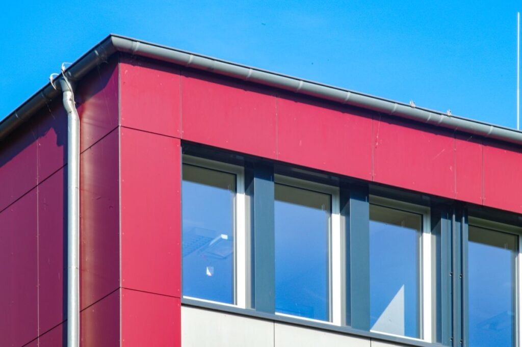 gutter materials A close-up view of the corner of a modern building with red exterior panels, large windows with reflections, and a gray gutter against a clear blue sky.