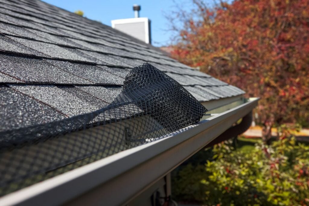 a gutter screens close-up of a house roof shows a section of black mesh gutter guard partially installed in the white rain gutter, with autumn trees blurred in the background.