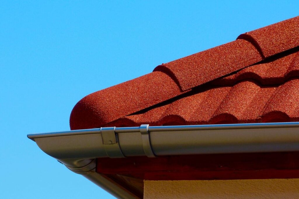 half round gutters Close-up of a house roof with red, curved tiles and a silver metal gutter against a clear blue sky.