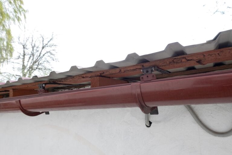 half round gutters Close-up of a red rain gutter attached to the edge of a corrugated metal roof on a white building. The sky is overcast and tree branches are visible in the background.