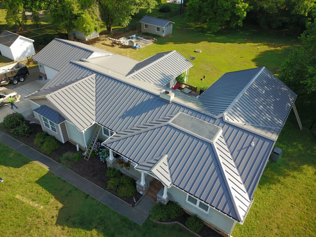 metal roof leak repair sealant Aerial view of a house with a gray metal roof, surrounded by grass and trees. The house has multiple roof angles and sections, with a driveway, garden, and outbuildings visible in the background.