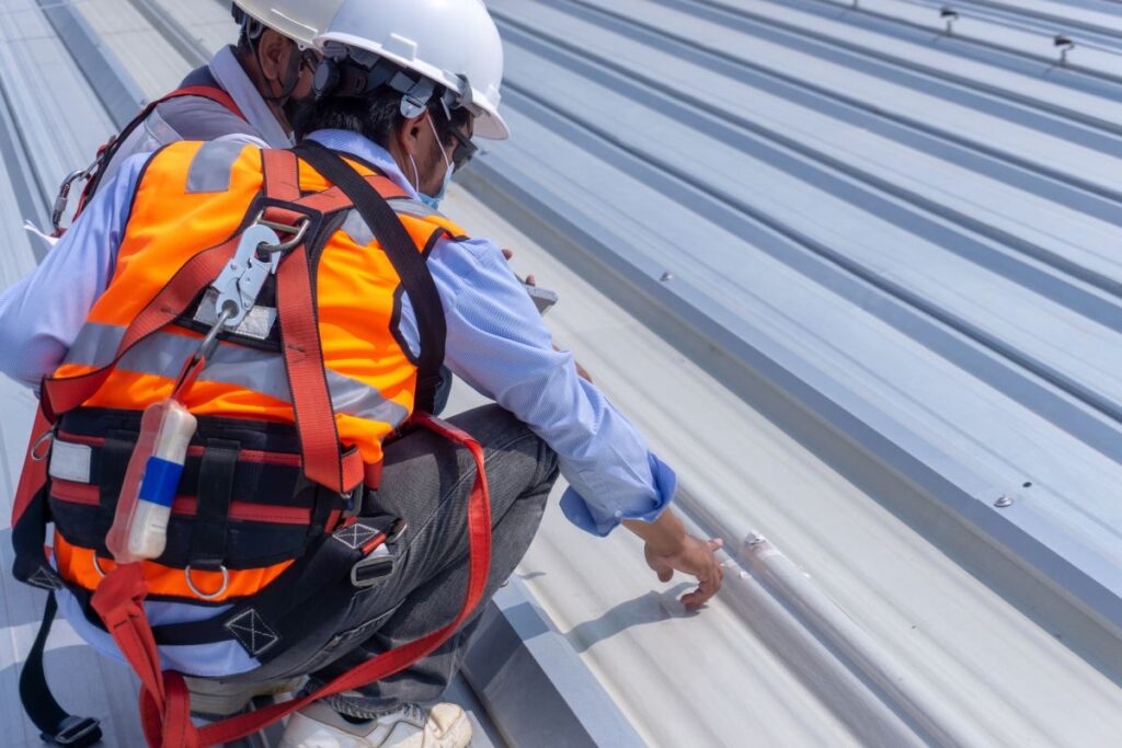 metal roof leak repair sealant Two workers in safety vests and helmets inspect a metal rooftop closely. One points at a specific spot on the roof, wearing a harness for safety. The bright metal roof panels are arranged in parallel lines.