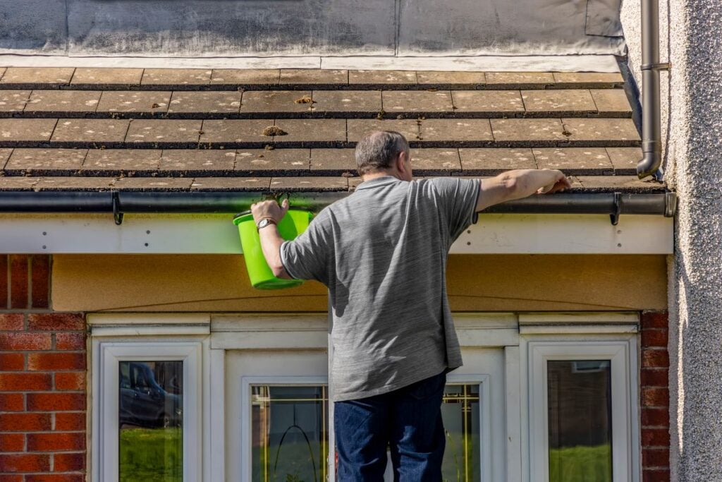 half round gutters A man in a gray shirt stands on a ladder cleaning leaves from a house gutter with his right hand, holding a green bucket in his left hand, above white-framed windows.