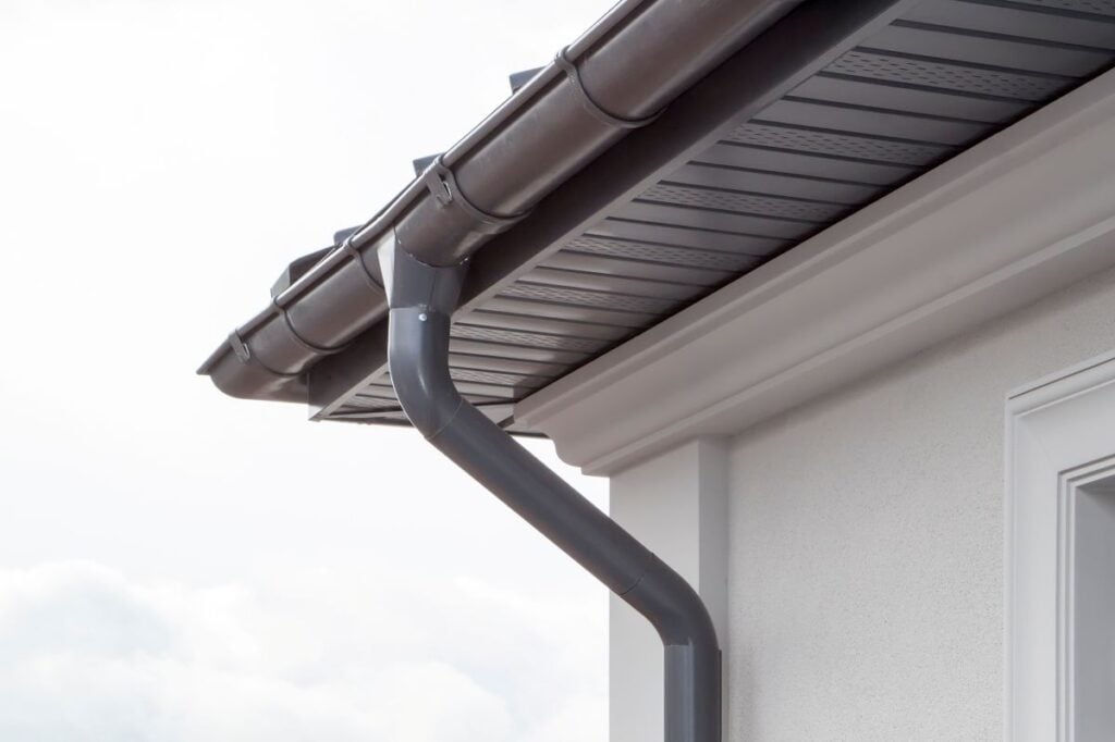 seamless gutters Close-up of a house roof corner showing a dark-colored rain gutter and downspout attached to the fascia, with part of the soffit and exterior wall visible against a cloudy sky.