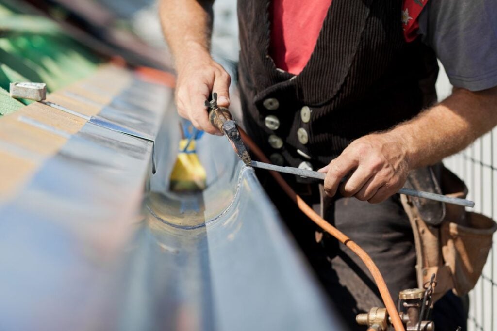 k style gutters close-up of a person wearing work clothes using a tool to weld or solder a metal surface outdoors, focusing on their hands and equipment.