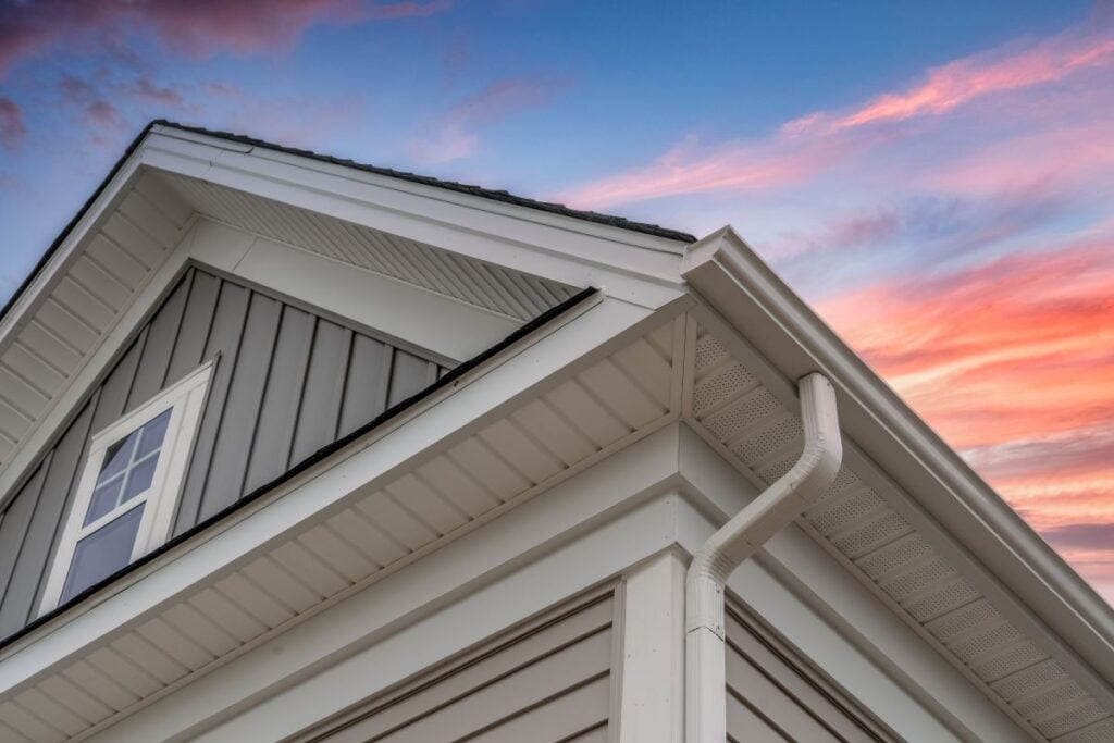 k style gutters Close-up view of a house's roof and rain gutter, with white siding and gray vertical panels, against a colorful sunset sky with pink and blue clouds.